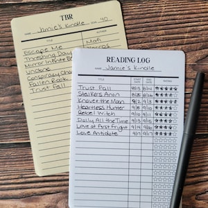 May include: A cream-colored TBR list card and a white Reading Log card, both with handwritten entries for Jamie's Kindle. The Reading Log includes columns for title, start date, end date, and star ratings. A black pen rests on the white card.