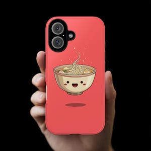 Puede incluir: Funda de teléfono roja con una ilustración de dibujos animados de un cuenco de ramen sonriente. El cuenco beige contiene fideos y caldo. La funda tiene una cámara dual y está en una mano.
