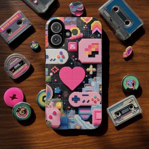 May include: A phone case with a pixelated design featuring a pink heart, a game controller, and other retro gaming elements. The case is surrounded by cassette tapes and colorful pins on a wooden surface.