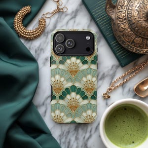 May include: A smartphone case with a green, white, and gold Art Deco fan pattern. The case is on a marble surface, with a gold necklace, a cup of green tea, and a decorative teapot in the background.