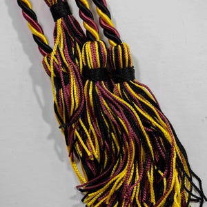 May include: Close-up of several decorative tassels. Each tassel features a black, yellow, and maroon colour scheme. The tassels are made of twisted cords and are likely used for graduation or decorative purposes.