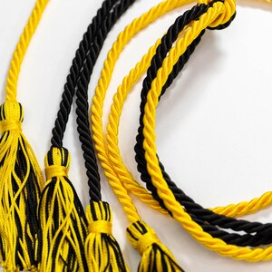 May include: Close-up of graduation cords in black and yellow. The cords are twisted and have tassels at the ends. The background is white.