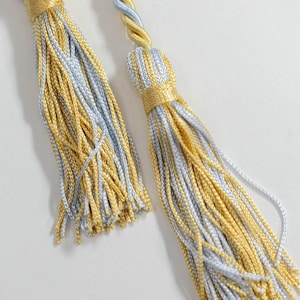 May include: Close-up of two decorative tassels. Each tassel features a combination of gold and silver-gray threads, with a twisted cord in matching colors at the top. The tassels are tied with gold thread, creating a luxurious and elegant look. Ideal for home decor.