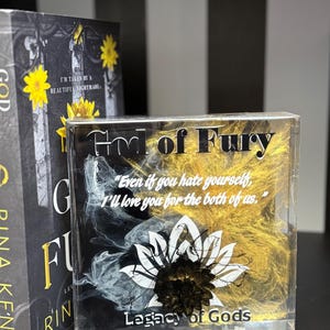May include: A clear resin block with the text "God of Fury" and "Legacy of Gods". The block features a black and white floral design with gold accents and a quote. A book with a yellow flower is in the background.