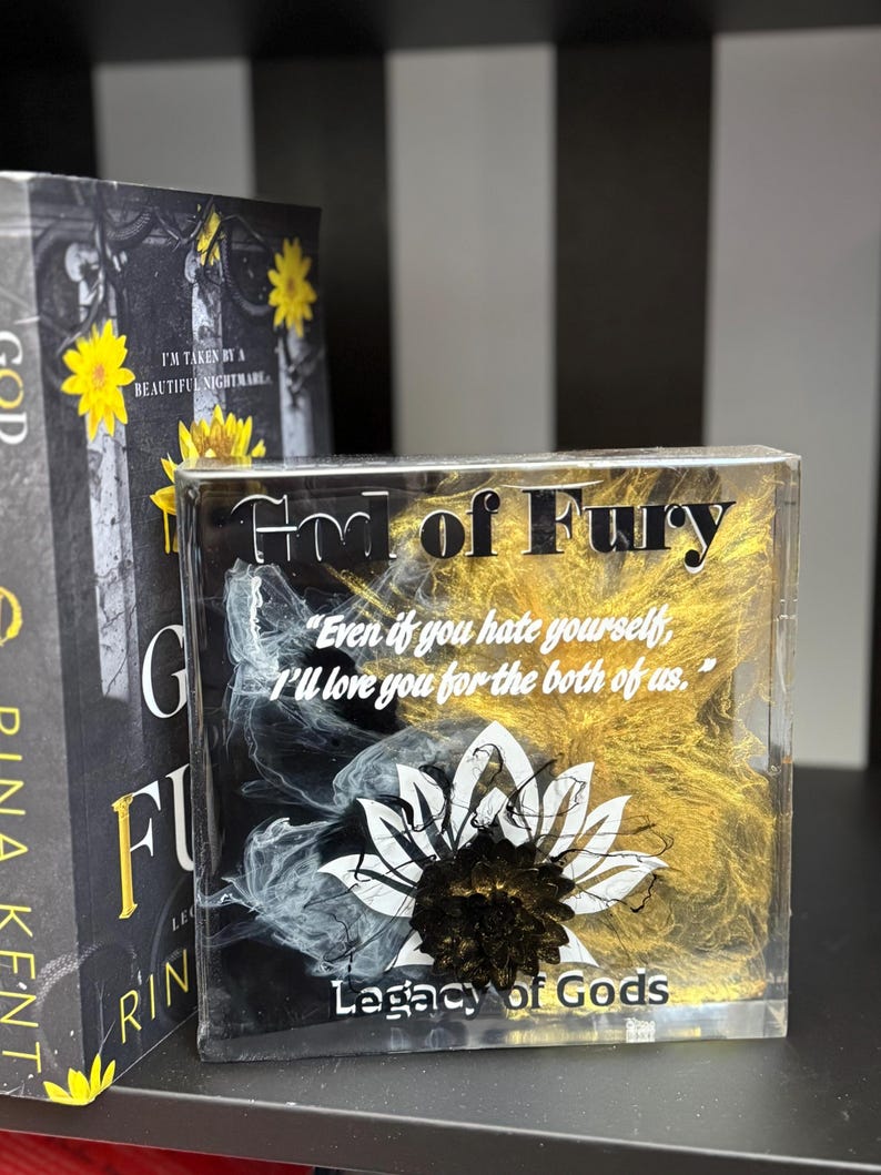 May include: A clear resin block with the text "God of Fury" and "Legacy of Gods". The block features a black and white floral design with gold accents and a quote. A book with a yellow flower is in the background.