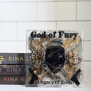 May include: A clear resin block with the text "God of Fury" and "Legacy of Gods". The block features a black mask, two guns, and floral designs. Three books with the author's name "Rina" are stacked to the left.