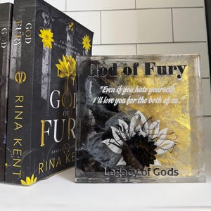 May include: A stack of books and a decorative resin block. The books have black covers with gold lettering and yellow flower accents. The resin block features the text "God of Fury" and a sunflower design with black, white, and gold accents.