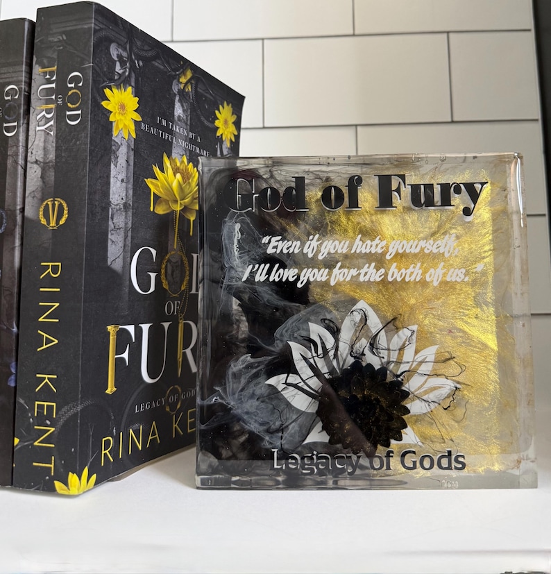 May include: A stack of books and a decorative resin block. The books have black covers with gold lettering and yellow flower accents. The resin block features the text "God of Fury" and a sunflower design with black, white, and gold accents.