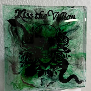 Puede incluir: Pieza de arte de resina cuadrada con un dise&ntilde;o verde y negro. La obra presenta una calavera negra, rosas y una serpiente, con las palabras "Kiss the Villain" en fuente cursiva. El fondo tiene un efecto verde veteado.