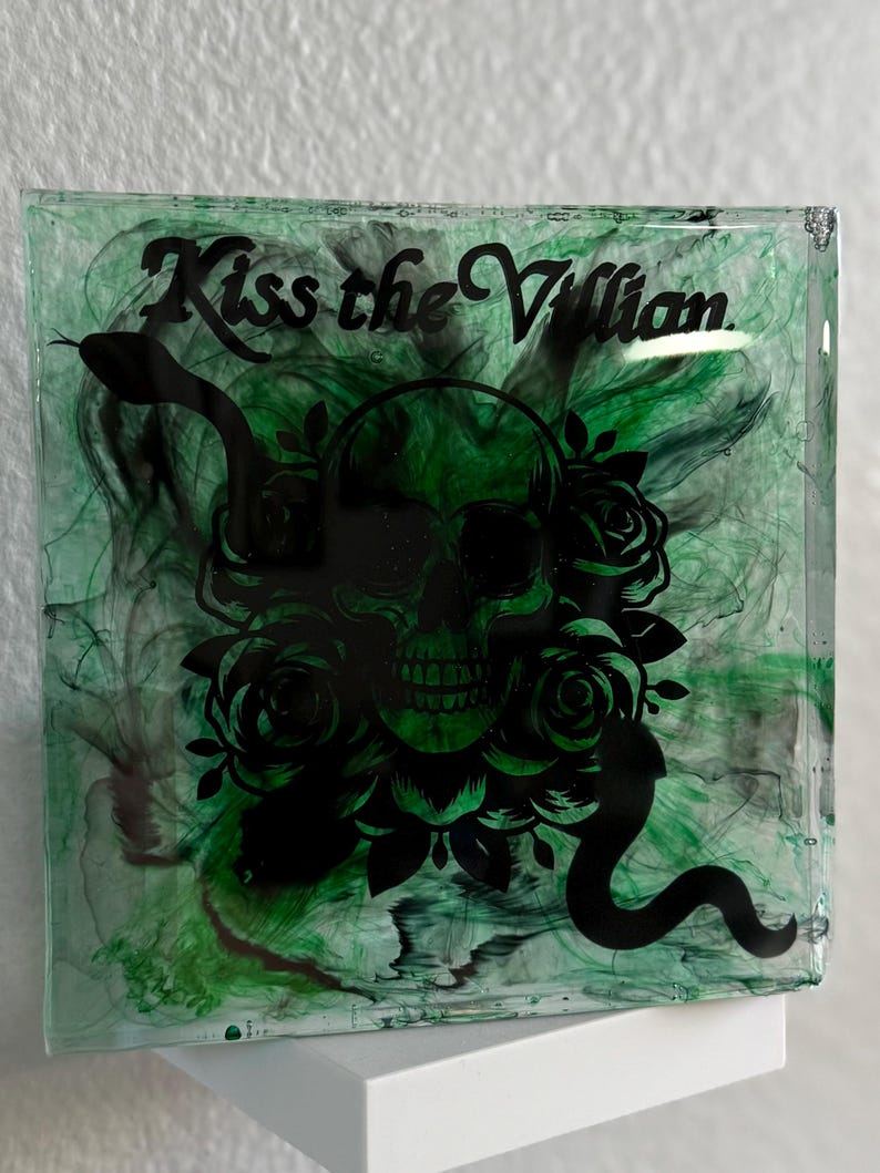 Puede incluir: Pieza de arte de resina cuadrada con un dise&ntilde;o verde y negro. La obra presenta una calavera negra, rosas y una serpiente, con las palabras "Kiss the Villain" en fuente cursiva. El fondo tiene un efecto verde veteado.