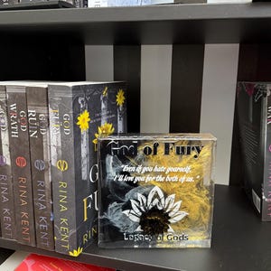 May include: A shelf displays several books from the "Legacy of Gods" series by Rina Kent. The book "God of Fury" is featured, along with a decorative block with the same title and a quote. The books have black spines with white text.