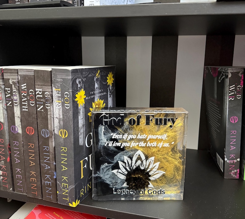 May include: A shelf displays several books from the "Legacy of Gods" series by Rina Kent. The book "God of Fury" is featured, along with a decorative block with the same title and a quote. The books have black spines with white text.