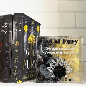 May include: A collection of books and a decorative block featuring the title "God of Fury" by Rina Kent. The books have black covers with gold and yellow accents. The block has the same title, a sunflower design, and a quote.