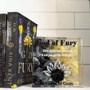 May include: A stack of books and a decorative block featuring the title "God of Fury" by Rina Kent. The books have black covers with gold lettering and a yellow flower design. The block has the same title, a quote, and a floral design.