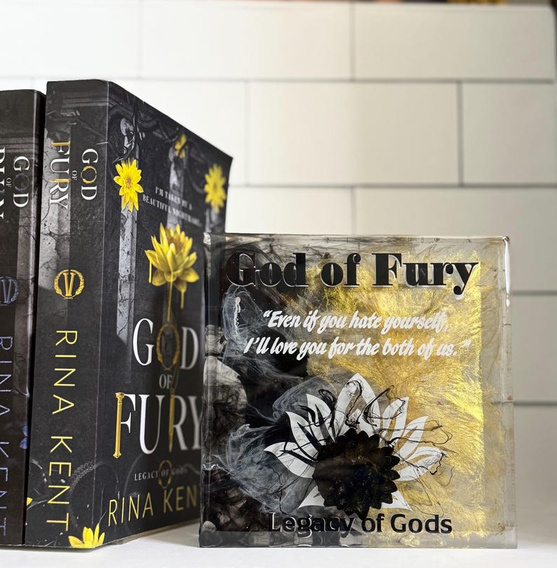 May include: A stack of books and a decorative block featuring the title "God of Fury" by Rina Kent. The books have black covers with gold lettering and a yellow flower design. The block has the same title, a quote, and a floral design.