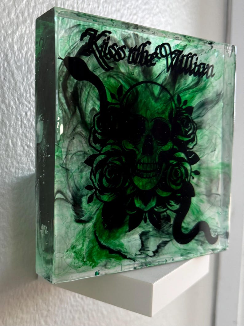 Puede incluir: Una pieza de arte de resina cuadrada con un fondo arremolinado verde y negro. La obra presenta una calavera negra con rosas, una serpiente y el texto "Kiss the Villain". La pieza se exhibe en un estante blanco.