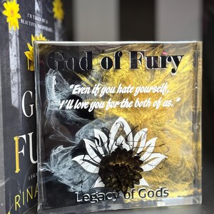 May include: A clear, rectangular block with the words "God of Fury" in black, gold, and white. The block features a white and black floral design and the text "Legacy of Gods". The quote "Even if you hate yourself, I'll love you for the both of us" is also visible.