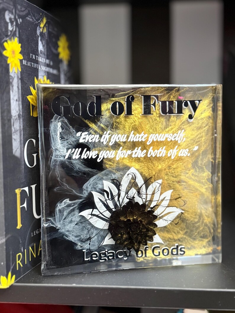 May include: A clear, rectangular block with the words "God of Fury" in black, gold, and white. The block features a white and black floral design and the text "Legacy of Gods". The quote "Even if you hate yourself, I'll love you for the both of us" is also visible.