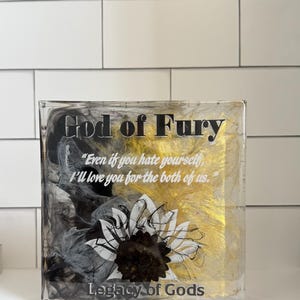 May include: A clear, rectangular block with the words "God of Fury" in black and gold. The block features a sunflower design and the text "Even if you hate yourself, I'll love you for the both of us." and "Legacy of Gods."