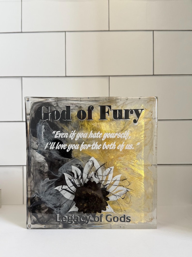 May include: A clear, rectangular block with the words "God of Fury" in black and gold. The block features a sunflower design and the text "Even if you hate yourself, I'll love you for the both of us." and "Legacy of Gods."