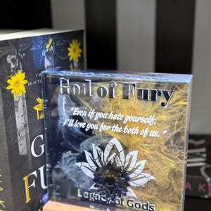 May include: A clear, rectangular resin block with the text "Heir of Fury" and a quote. The design features a white lotus flower and gold and black accents. It sits on a wooden base.