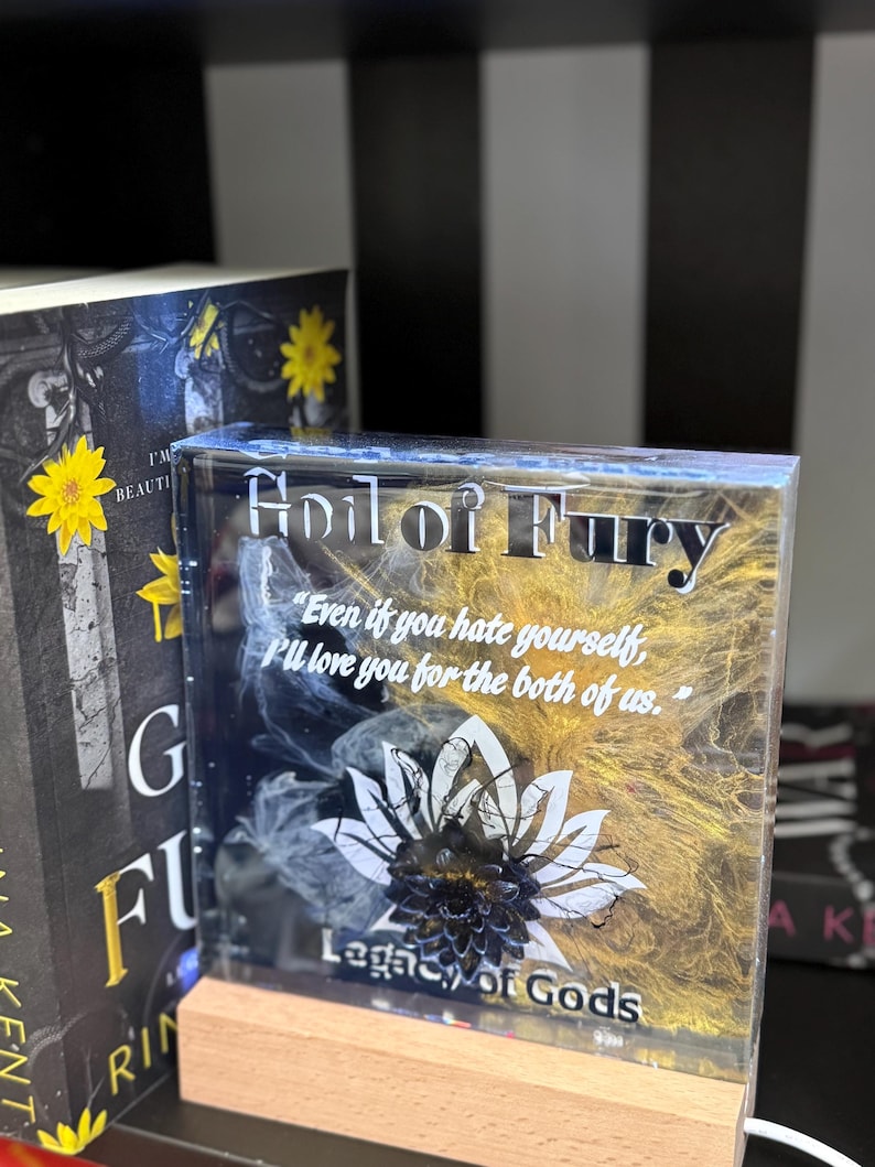 May include: A clear, rectangular resin block with the text "Heir of Fury" and a quote. The design features a white lotus flower and gold and black accents. It sits on a wooden base.
