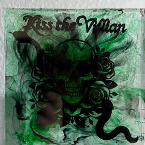 Puede incluir: Posavasos cuadrado con un dise&ntilde;o en espiral verde y negro. Una calavera negra con rosas y una serpiente est&aacute; centrada, con el texto "Kiss the Villain" arriba. El posavasos est&aacute; hecho de un material transparente y brillante.