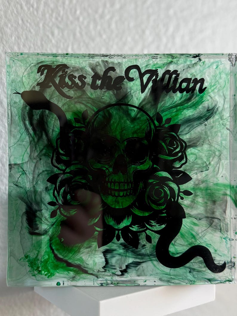 Puede incluir: Posavasos cuadrado con un dise&ntilde;o en espiral verde y negro. Una calavera negra con rosas y una serpiente est&aacute; centrada, con el texto "Kiss the Villain" arriba. El posavasos est&aacute; hecho de un material transparente y brillante.