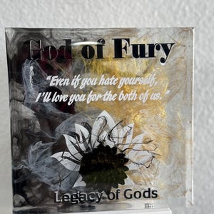 May include: A square, clear resin art piece with the text "God of Fury" and "Legacy of Gods." The artwork features a white and black floral design, gold accents, and a quote. It is mounted on a white base.