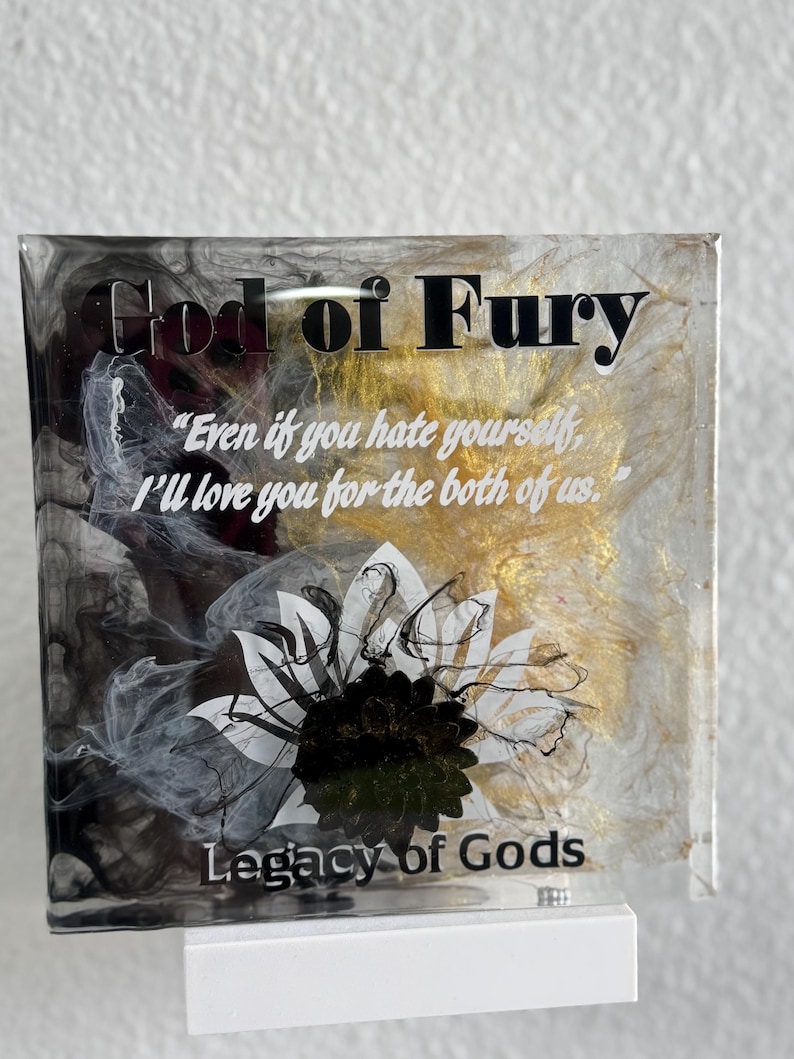 May include: A square, clear resin art piece with the text "God of Fury" and "Legacy of Gods." The artwork features a white and black floral design, gold accents, and a quote. It is mounted on a white base.