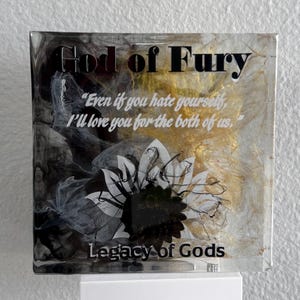 May include: A square, clear resin art piece with the text "God of Fury" and a quote in white. The background features black, gold, and white abstract designs, with a floral graphic. The base is white, and the text "Legacy of Gods" is at the bottom.