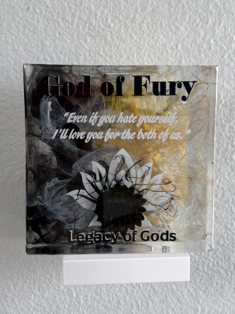 May include: A square, clear resin art piece with the text "God of Fury" and a quote in white. The background features black, gold, and white abstract designs, with a floral graphic. The base is white, and the text "Legacy of Gods" is at the bottom.