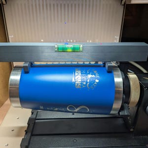 May include: A blue tumbler with silver text and a graphic is held in a black machine. A level is placed on top of the tumbler. The tumbler is being prepared for engraving.