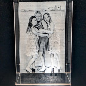 May include: A rectangular crystal photo block featuring an etched image of three people embracing. The image is in grayscale, showing the figures in dresses. The crystal block is clear and stands upright.