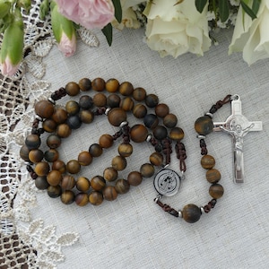 May include: A rosary featuring matte brown tiger eye beads strung on dark brown cord. The rosary includes a circular silver-toned metal centerpiece with an embossed design and a silver-toned metal crucifix featuring a figure of Jesus.