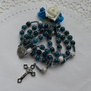 May include: A rosary with teal and white beads, a silver crucifix, and a medal. A white and blue floral accent sits at the top. The rosary is displayed on a white, patterned surface.