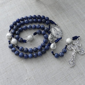 May include: A rosary with blue and white beads. The rosary features a silver crucifix and a silver pendant with an image. The beads are a mix of dark blue and white, with a textured appearance.
