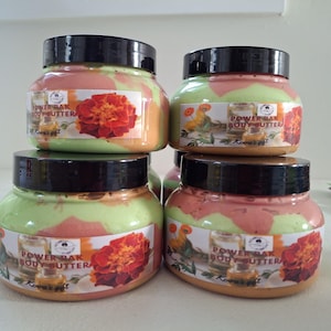 May include: Four jars of Power Bak Body Butter are stacked. The jars have black lids and labels with a marigold flower and text. The body butter inside is a mix of pink, orange, and green colours.