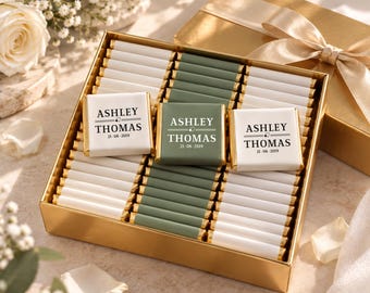 May include: A gold gift box filled with rows of small chocolate bars wrapped in white and sage green paper. Three bars feature the printed text ASHLEY THOMAS 21-08-2019 with a small heart icon. The box is topped with a matching gold ribbon.