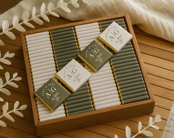 May include: A wooden tray filled with individually wrapped chocolates. The chocolates are arranged in rows of white and olive green. Four chocolates on top have the initials "A/G" and floral designs.