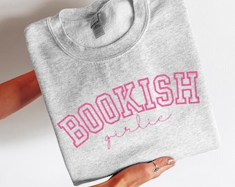 May include: A heather gray sweatshirt with the text "BOOKISH girlie" in pink. The sweatshirt is held by a person. The text is in a collegiate style font.