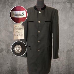 Puede incluir: Una chaqueta gris oscuro de manga larga con ribetes de terciopelo negro y botones dorados. La chaqueta tiene un cuello alto con ribete rojo. Una etiqueta dice "Nockstein Trachten" y enumera la composición de la tela. La chaqueta se exhibe en un maniquí.