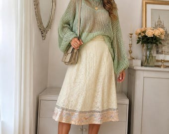 May include: A sage green loose-knit long-sleeve sweater with a wide neckline, paired with a cream-colored midi skirt featuring a delicate lace overlay and a decorative hem band with subtle floral embroidery, worn with beige loafers and a shoulder bag.