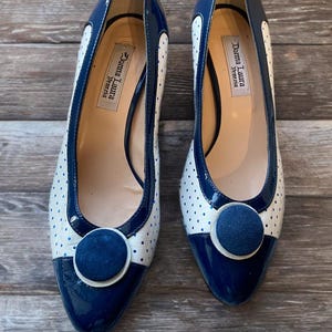 May include: Pair of navy blue and white polka dot heels with a pointed toe and a round navy blue button detail. The heels have a patent leather finish and the brand name "Donna Laura Venezia" is visible inside.