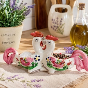 Puede incluir: Un servidor de condimentos doble de cerámica con forma de dos gallos mirando en direcciones opuestas. La pieza es de color crema con plumas de cola rosas, follaje verde y pequeñas flores azules y amarillas pintadas a mano en cada lado.