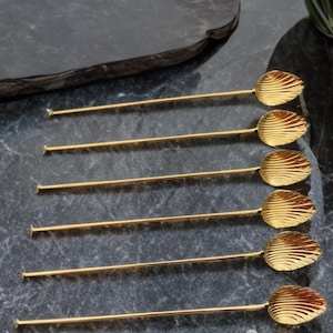 Puede incluir: Seis cucharas doradas para servir con un diseño en forma de hoja en la cabeza de la cuchara. Las cucharas tienen mangos largos y delgados y están dispuestas sobre una superficie oscura y texturizada. Las cucharas son probablemente para servir aperitivos o postres.