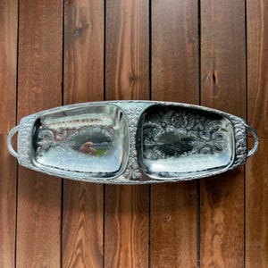 Puede incluir: Bandeja de servir plateada de dos secciones con detalles florales y de volutas ornamentados. La bandeja tiene forma rectangular con extremos redondeados y asas a cada lado. El fondo es una superficie de madera.