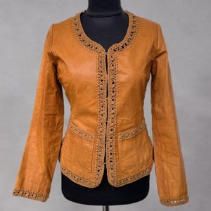May include: A tan leather jacket with a round neckline and long sleeves. The jacket features decorative crochet trim along the collar, front opening, pockets, and cuffs. The jacket is displayed on a black mannequin.