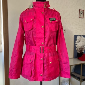 May include: A bright pink jacket featuring a high collar with a buckle strap, a front button closure, and a matching waist belt with a gold-tone buckle. The jacket includes four front flap pockets with gold-tone buttons and a Barbour International patch.