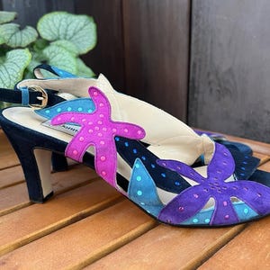 Puede incluir: Sandalias de tacón de ante negro con recortes de estrellas de mar y lunares de colores. Las sandalias tienen una correa con hebilla dorada y tacón alto. Las estrellas de mar son rosas y moradas, y los lunares son azules.